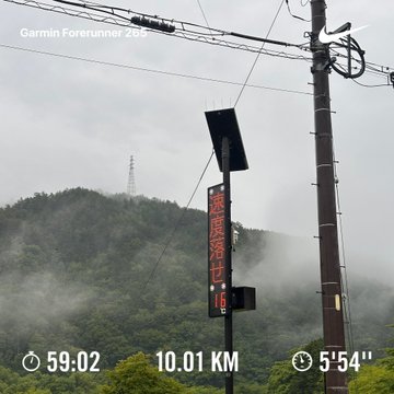 The image shows a street sign with a red neon number "16" displayed, situated next to an electric pole. The background includes mountains covered in fog, giving the scene a cool and mysterious atmosphere. The location appears to be a forested area near a mountain range, possibly at the foot of a volcano or another natural formation. The image has been taken with the step numbers 5o9-02 and 100x3 written on it, indicating that this was captured during a run through the forested area.