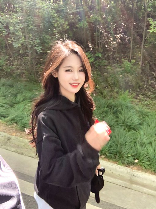 A photo of a young woman with long dark hair wearing a black hoodie, smiling at the camera in an outdoor setting with green foliage behind her.