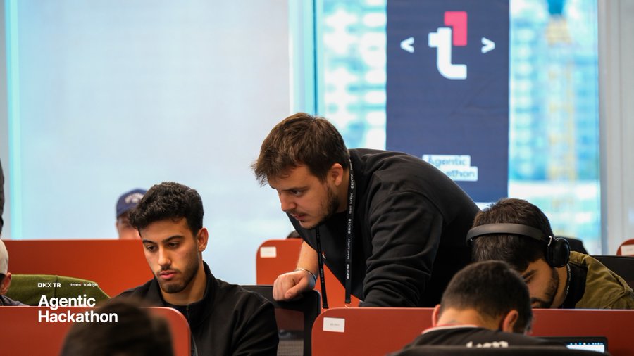 Young men working intently on laptops at the 'Agentic Hackathon' sponsored by DK and TR, with a large illuminated 't' logo in the background of what appears to be a modern office or event space.