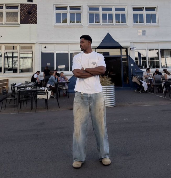 A young man stands with crossed arms on a sidewalk in front of a white building, wearing a white t-shirt, light wash baggy jeans, and tan sneakers.