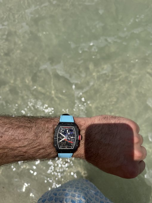 The image features a man's wrist with a watch, which has a blue strap. The watch appears to be black and white or silver, depending on the lighting. The background shows water that looks like it could be a lake or ocean, suggesting that the photo was taken at the beach or near the shoreline. The caption of the image reads "Be blue in a sea of red Hyperliquid", which is likely an artistic or creative statement about the colors and atmosphere surrounding the man's watch.