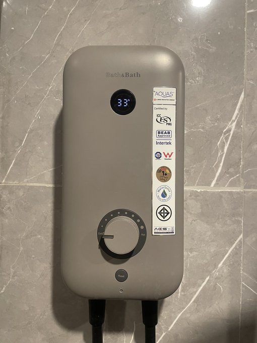 A wall-mounted Becha Bath electric water heater displaying a temperature of 33 degrees on a digital screen, with various certification labels and a control dial.