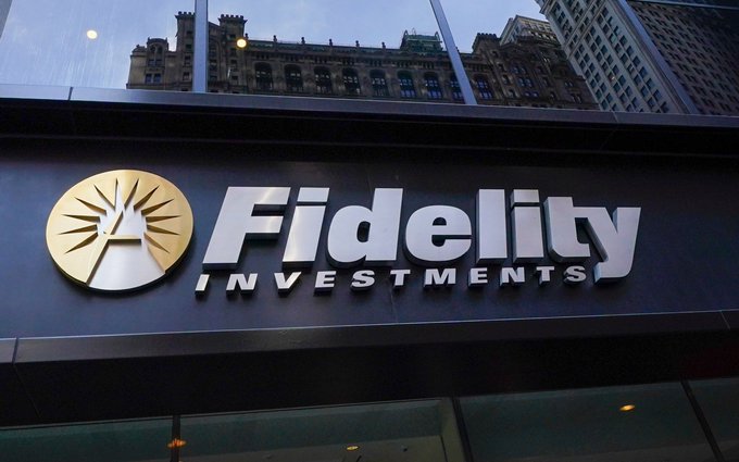 The image shows a Fidelity Investments storefront sign with the company's gold sunburst logo and bold white lettering against a dark background, with city buildings reflected above. The image visually represents Fidelity as a company, directly connecting to the post about Fidelity's plans to add staking to its Ethereum ETF (FETH) and distribute rewards to investors.