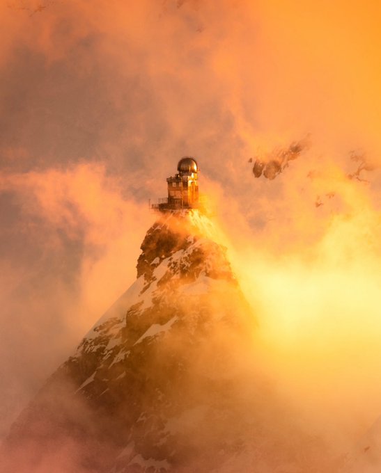 A dramatic scene of a snow-capped mountain peak topped with a domed observatory building, illuminated by golden sunlight breaking through thick orange-tinted clouds.