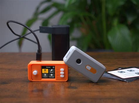 A photograph of two small electronic devices, one orange and one gray, sitting on a wooden table with a plant in the background; these appear to be Bitcoin/crypto hardware wallets.