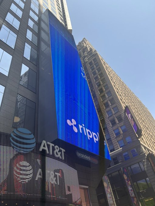 A massive billboard displaying Ripple's logo and name on a bright blue digital screen is mounted on a building alongside AT&T signage in a bustling city setting. The advertisement aligns with the post celebrating Ripple's prominent new ad visible in New York City.