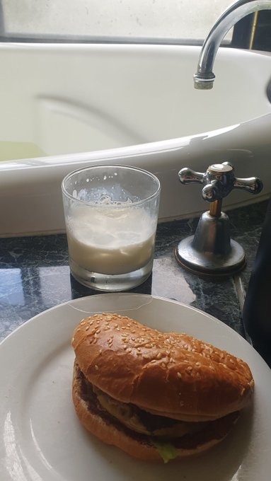 A simple meal consisting of a sesame seed bun burger on a white plate next to a glass of milky beverage, placed on a bathroom counter beside a bathtub.