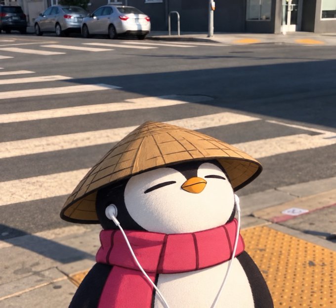 A plush penguin wearing a traditional conical straw hat, a pink scarf, and white earphones stands smiling at a crosswalk.