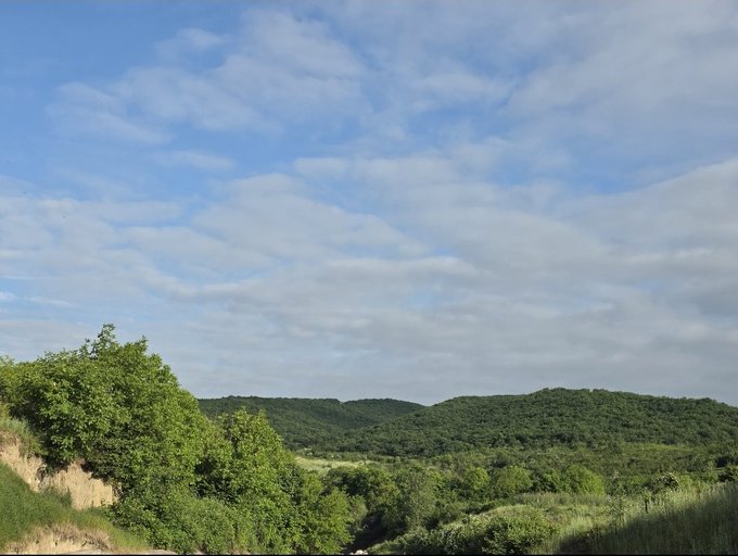 A scenic landscape view shows rolling green forested hills under a bright blue sky with scattered white clouds.