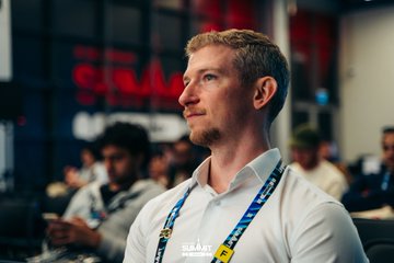 A man in a white shirt with a lanyard looks off to the side at what appears to be a conference or event, with other attendees visible in the blurred background.
