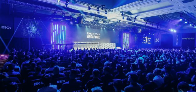 A large crowd gathers at the 'TOKEN2049 SINGAPORE' conference in a hall bathed in blue stage lighting, with 'OKX' logos visible around the venue.