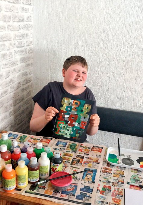 A smiling young boy sitting at a table covered with newspaper, surrounded by numerous bottles of colorful acrylic paints and painting supplies.