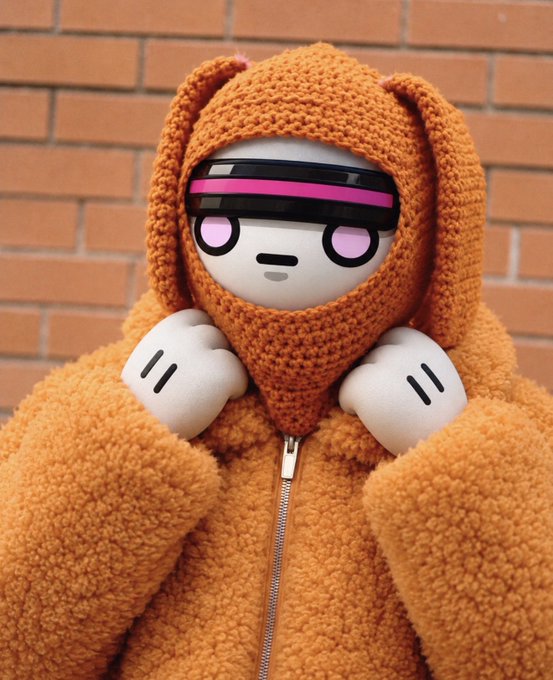 A figure or doll with a white round face, black and pink visor, and white paws wears a bulky orange knit hooded jacket against a brick wall background.