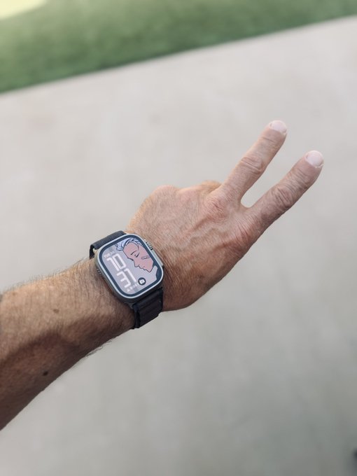 A person's outstretched arm displays a black smartwatch with a black band; the watch face features a cartoon cloud graphic and reads '12:05' against a backdrop of concrete and grass.