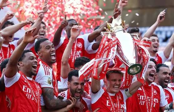 The image shows a group of people celebrating their victory at a soccer match, with two men holding up a trophy. The trophy has a gold crown on top, indicating that they have won the Champions League. The players are all wearing red jerseys, which is typical for soccer teams. They are standing in front of a building and there are confetti falling from above, adding to the festive atmosphere.