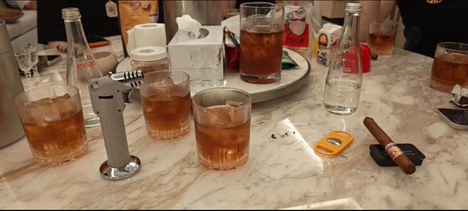 The image shows a table with several glasses filled with drinks, including beer and cocktails such as martinis and champagne. There are also some snacks and a cigar on the table. The scene appears to be set for a social gathering or celebration among friends or colleagues. In the background, there is a person who seems to have left the room, possibly after the event has ended. The post about JU Coin A group fleeing to Singapore suggests that this gathering might have been related to an illegal activity or some form of financial transaction involving the JU Coin A group.