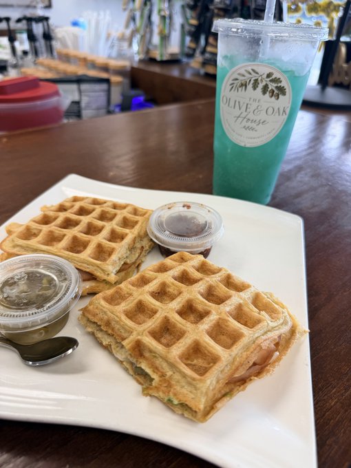 A brunch plate with two Belgian-style waffles served with small cups of syrup and jam, accompanied by a blue drink from 'The Olive & Oak House' café.