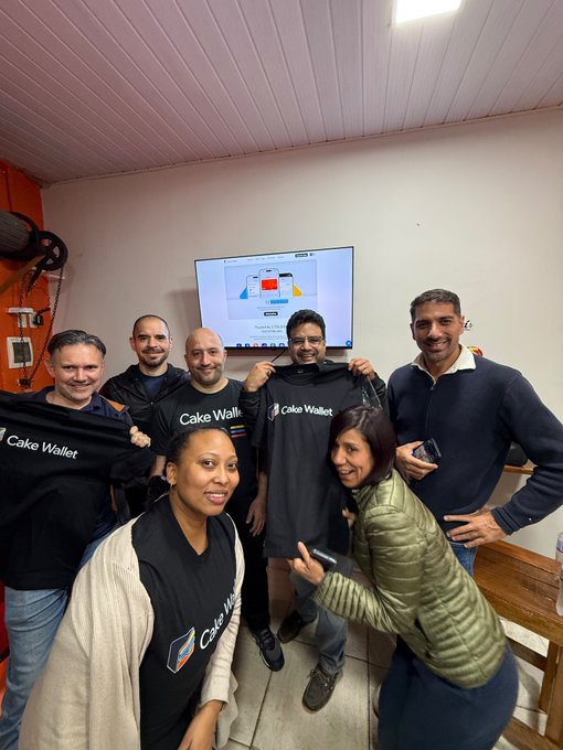 A group of smiling people pose for a photo indoors, with several wearing or holding black t-shirts that read 'Cake Wallet'.