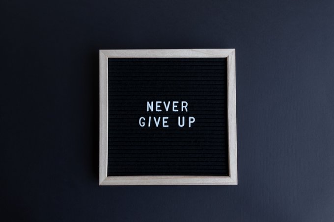 A minimalist black letter board in a light wooden frame mounted on a dark wall, displaying the motivational message "NEVER GIVE UP" in white letters.