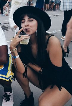 A woman wearing a black hat and black outfit crouches while drinking from a clear plastic cup at an outdoor event.