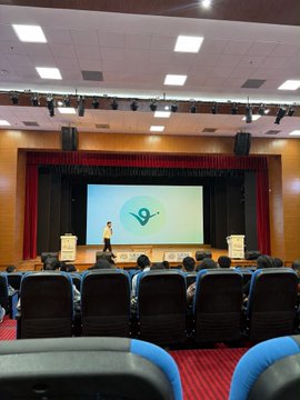 A speaker presents on stage at a Robinhood Crypto x Virtuals builder session in Delhi, with a large screen displaying the "V" Virtuals logo to an audience seated in an auditorium. The event introduces students to the Virtuals.io platform and Robinhood Crypto Chain, showcasing the collaborative educational initiative in India.