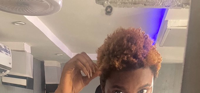 A selfie of a person with curly blonde hair in a bathroom, looking up at the camera while touching their hair.
