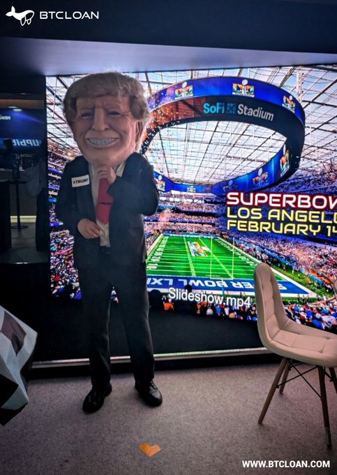 A person stands in front of a large screen showing a Super Bowl advertisement for SoFi Stadium, with text including 'SUPERBOW LOS ANGEL', 'FEBRUARY 14', 'Slideshow.mp4', and the BTCLOAN logo visible.