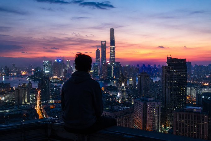 A silhouetted figure sits on a rooftop overlooking a sprawling city skyline at sunset, featuring illuminated skyscrapers.