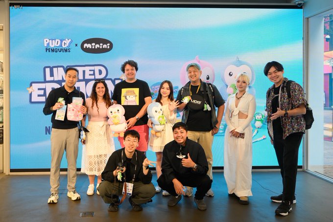 A group of eight people pose for a photo at a promotional event for 'Pudgy Penguins x minimini,' holding plush toys in front of a large blue screen displaying the collaboration's logo.