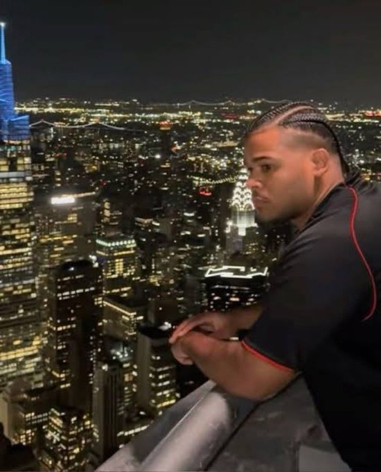 A man with braided hair perches on the edge of a high-rise rooftop, gazing contemplatively over a sprawling nighttime cityscape with One World Trade Center visible in the distance. The image humorously contrasts the vast, complex urban environment with the "memecoin" conversation—suggesting that while others are distracted by fleeting crypto trends, he sees the bigger, distant view (a nod to either artistic vision or simply tuning out the noise).