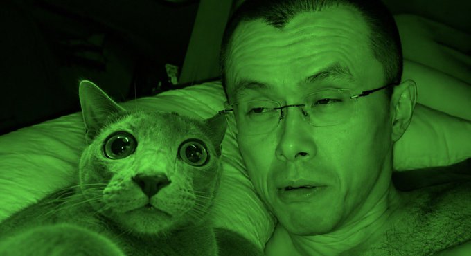 A man and a cat rest side-by-side in a green-tinted night vision image, both staring wide-eyed at the camera. The eerie green hue and the subjects' alert expressions suggest a quiet, late-night moment captured just before something is about to happen—fitting the simple "Soon" caption as a teaser for an impending moment.