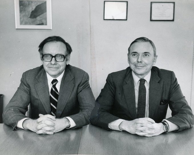 A black and white vintage photograph of two men in suits and ties sitting side by side at a desk with framed pictures on the wall behind them.