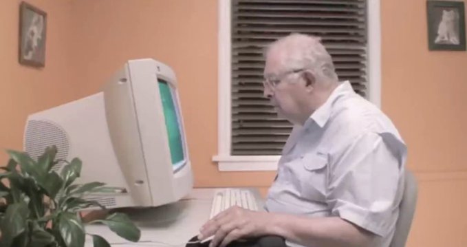 An older man with glasses wearing a light blue short-sleeved shirt sits at a desk using an old beige CRT computer monitor with a green screen, with a plant and window blinds visible in the background.
