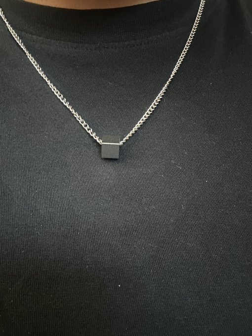 A close-up photograph of a silver chain necklace with a small rectangular pendant, worn against a black t-shirt.