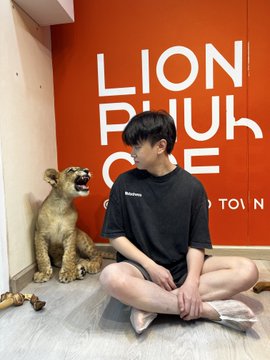 A young man sits cross-legged on the floor wearing a black t-shirt, positioned next to a taxidermied lion, with 'LION PHUKET' text visible on an orange wall behind them.