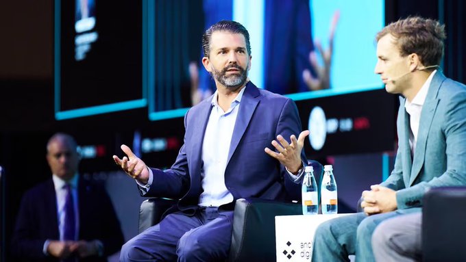 Donald Trump Jr. speaks with animated hand gestures while seated next to another man on a panel stage at what appears to be a Binance-related event, with water bottles in front of them.