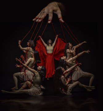 A giant hand controls several women like puppets using red strings against a black background. One woman in the center wears a red dress, while the others wear tan clothing.