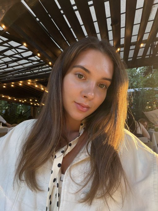 A selfie of a woman with long brown hair wearing a white shirt and a black-and-white polka dot scarf, taken under a pergola with string lights.