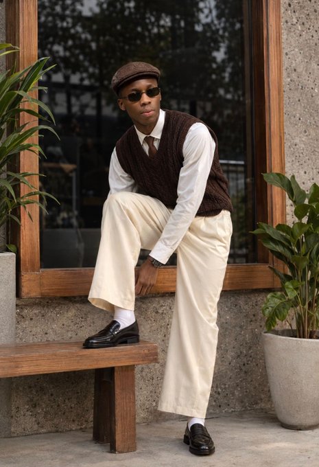 A stylish person poses on a wooden bench, wearing a brown knit vest over a white dress shirt, cream-colored trousers, dark loafers, and a flat cap with sunglasses, leaning casually against a window frame surrounded by green plants. The outfit's polished, vintage-inspired fashion fits well with a brand-promoting social media post by GM CT (@RiverdotInc, @River4fun).
