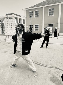 The image features a man dressed in all white performing a dance move with his arms outstretched while standing on a street. The photo has been edited to appear black and white, giving it an artistic touch. In the background, there are two people who seem to be observing or participating in the dance performance. The scene takes place outside of a building, possibly a store or a public space where such activities can occur.