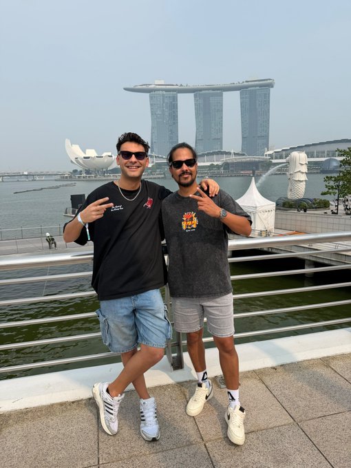 Two men posing together in front of the Marina Bay Sands hotel in Singapore, with one wearing a black t-shirt and the other in a grey elephant-print shirt, both making peace signs.