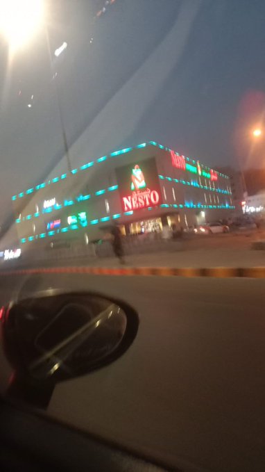 A blurry nighttime photo taken from inside a car, showing an illuminated building with signs for 'Nesto' and 'Alyasr'.