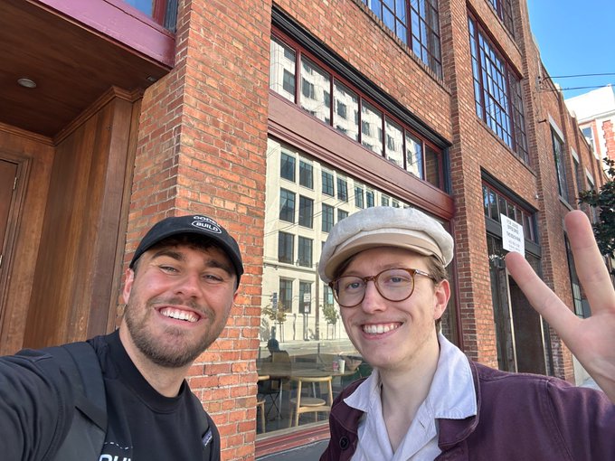 Two smiling men pose for a selfie in front of a brick building with large windows; one wears a black cap and the other wears a beige cap with glasses.