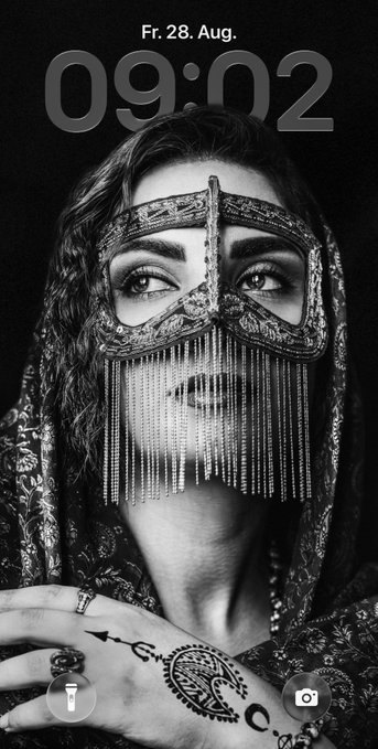 A black and white lock screen wallpaper showing a woman with an ornate beaded veil over her face and henna designs on her hand, displaying the time "09:02" and date "Fr. 28. Aug.".