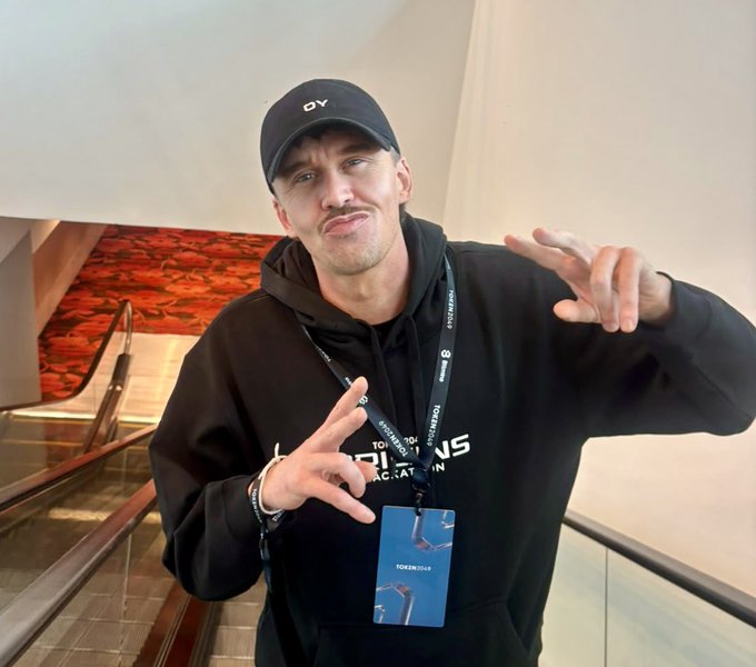 A man wearing a black hoodie and a black cap with 'CY' on it poses with hand gestures, wearing a lanyard with 'TOKEN2049' and 'PITCH STORM' text on the badge.