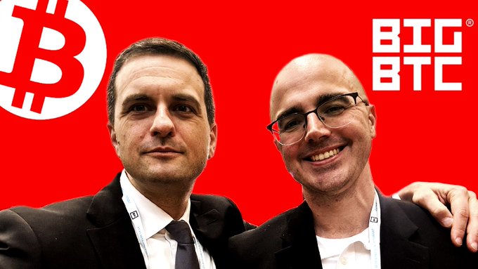 Two men posing together in front of a red Bitcoin background featuring the BTC logo and 'BTC BTC' text.
