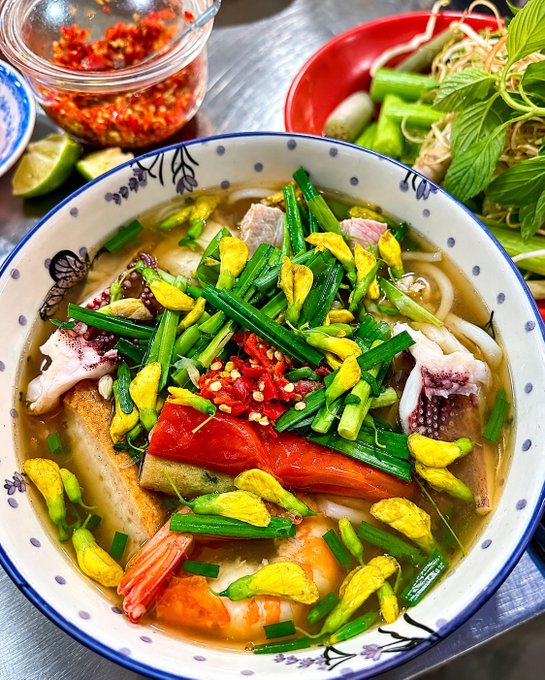 A bowl of Vietnamese soup with rice noodles, shrimp, squid, tomatoes, and abundant green onions and herbs, accompanied by chili sauce and lime on the side.