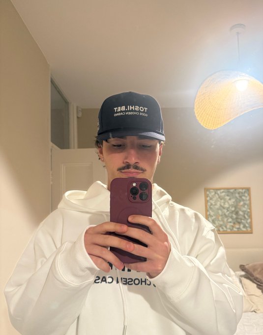 A young man takes a mirror selfie wearing a white "CHOOSE ICAS" hoodie and a navy cap reading "TOSHI.BET" and "ONLINE ESPORTS BONDS," reflecting the post's message about Toshi bet providing branded apparel to its affiliates. The dimly lit bedroom setting and casual pose emphasize the promotional merchandise aspect highlighted in the caption.