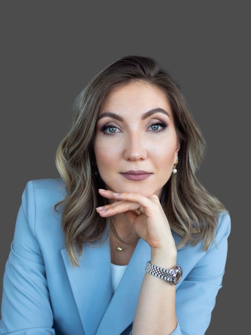 A professional headshot of a woman with brown hair wearing a light blue blazer, a watch, and a delicate necklace, resting her chin on her hand against a gray background.