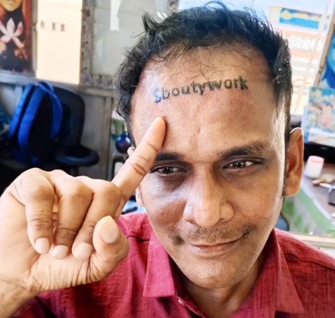 A photo of a man with a forehead tattoo reading "Showtywork" pointing his index finger at the text while wearing a red shirt in what appears to be a barbershop setting.